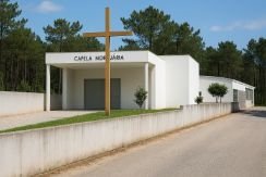 Vista Inspirada Na Casa Mortuária De Santana, Em Ferreira-a-nova, Com Edifício Branco, Cruz Frontal E Vegetação Envolvente, Representando Património Local.